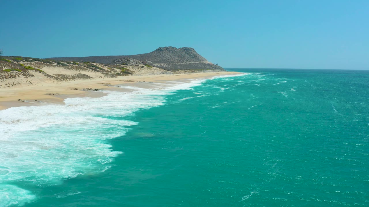 At La Laguna Beach Los Cabos, the surf curls across the left shoreline as turquoise water stretches to the right and dunes lead back toward a rocky hill, no visible people.