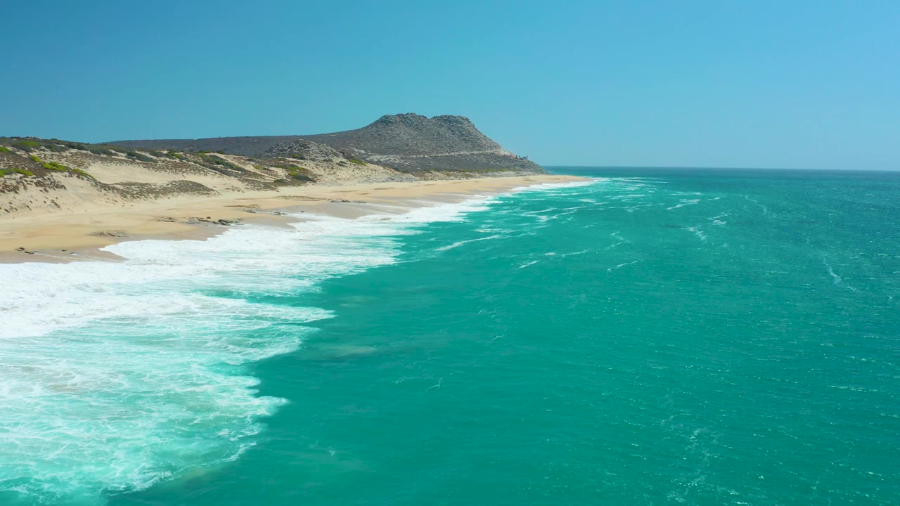 At La Laguna Beach Los Cabos, white water spreads over the lower left sand while the shoreline bends toward the distant hill and the open turquoise sea remains clear, no visible people.