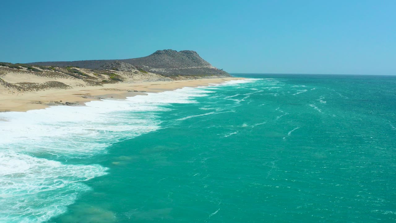 At La Laguna Beach Los Cabos, turquoise water and white surf run along the sandy shore with a rocky hill in the distance and no visible people.