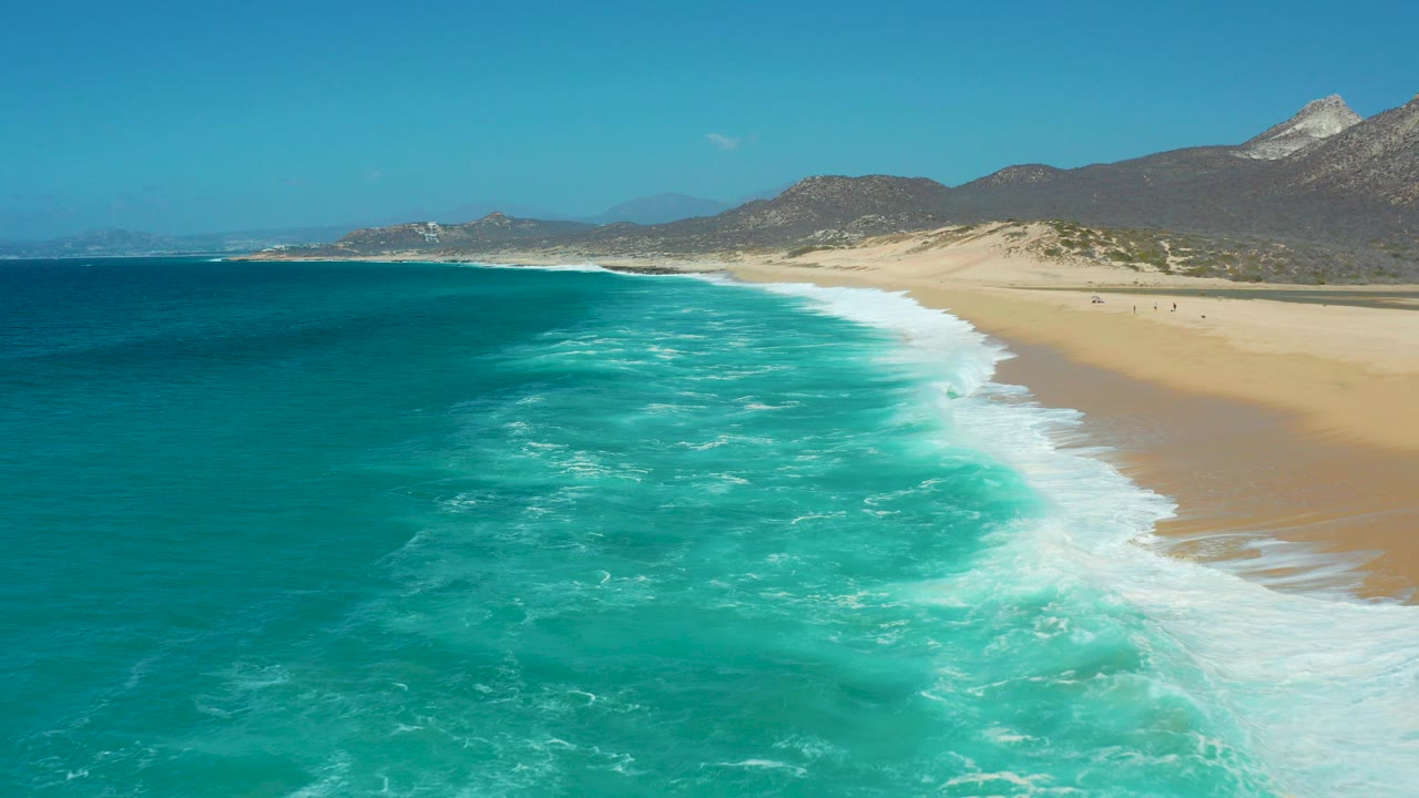Wide aerial view over La Laguna Beach in Los Cabos, with turquoise water in the foreground, white surf along the sand, distant mountains, clear sky, and no visible people.