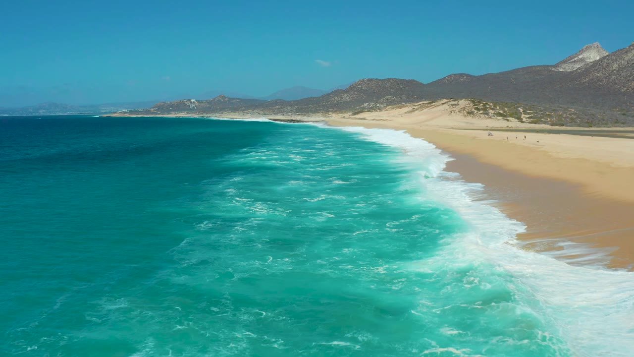 Aerial frame from La Laguna Beach in Los Cabos showing foamy turquoise water moving toward the right-side sand, with low dunes, mountains, clear sky, and no visible people.