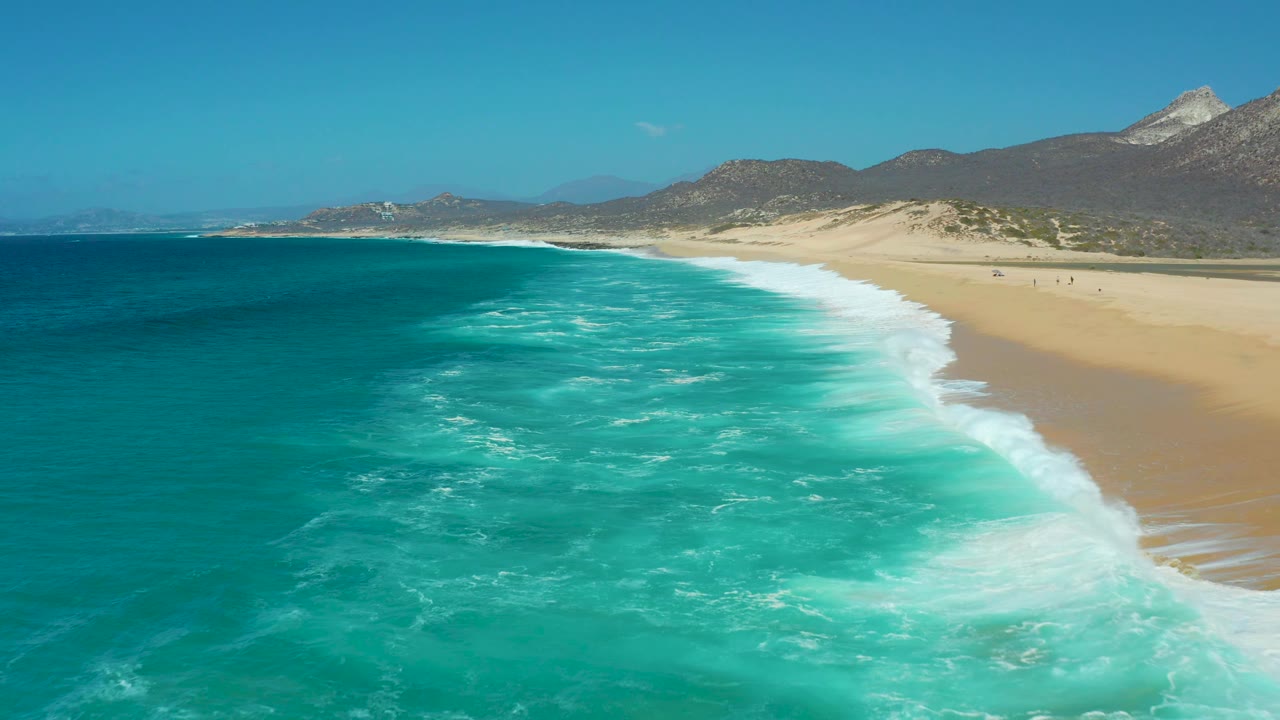 La Laguna Beach appears from above with open turquoise water across the foreground, a bright foam band at the shore, sand to the right, distant mountains, and no visible people.