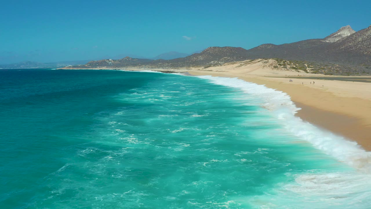 White surf curves along a wide sandy beach below dark coastal mountains.