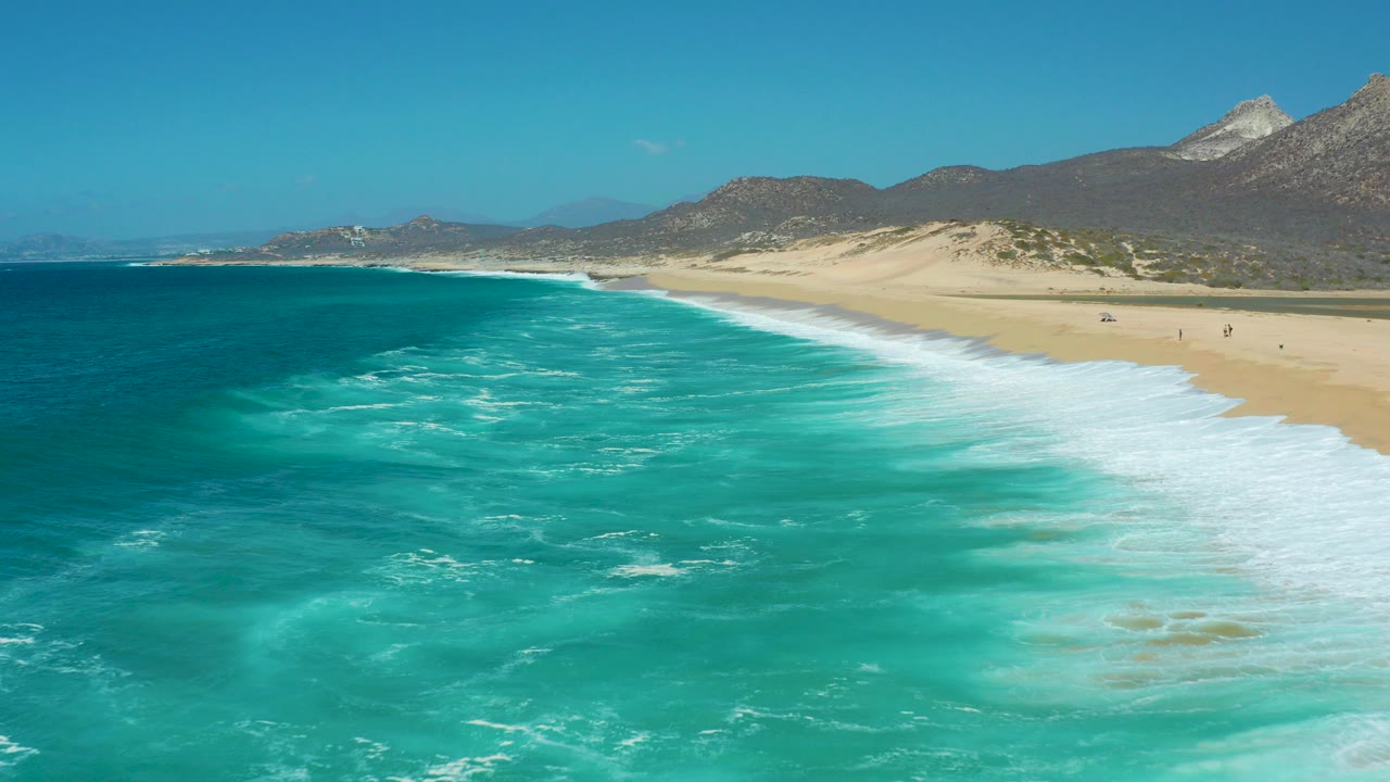 At La Laguna Beach Los Cabos, turquoise ocean and white surf angle toward a broad sandy shore with dunes and mountains beyond, with tiny distant beachgoers near the right side.