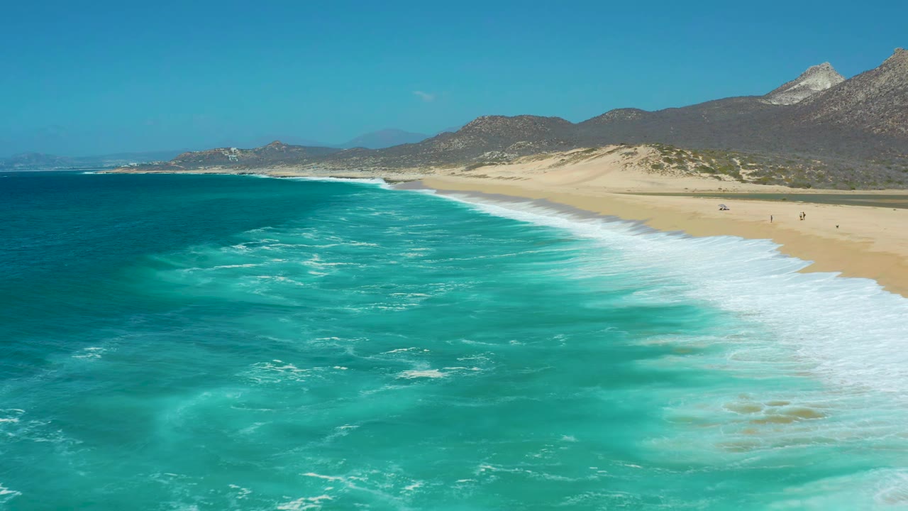 The La Laguna Beach Los Cabos shoreline stretches diagonally as foamy turquoise water moves toward pale sand, with dry hills, mountains, clear sky, and small distant figures on the beach.