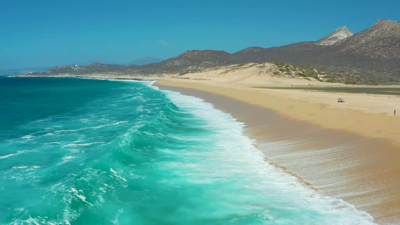 La Laguna Beach Los Cabos coastline seen from above as a larger band of white foam pushes toward tan sand, with dunes, mountains, blue sky, and no visible people.