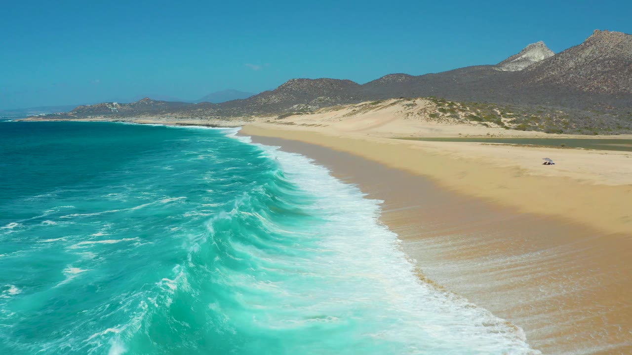 Elevated view over La Laguna Beach Los Cabos where the turquoise ocean curves along the left side and foam spreads across the shore, with dunes and no visible people.