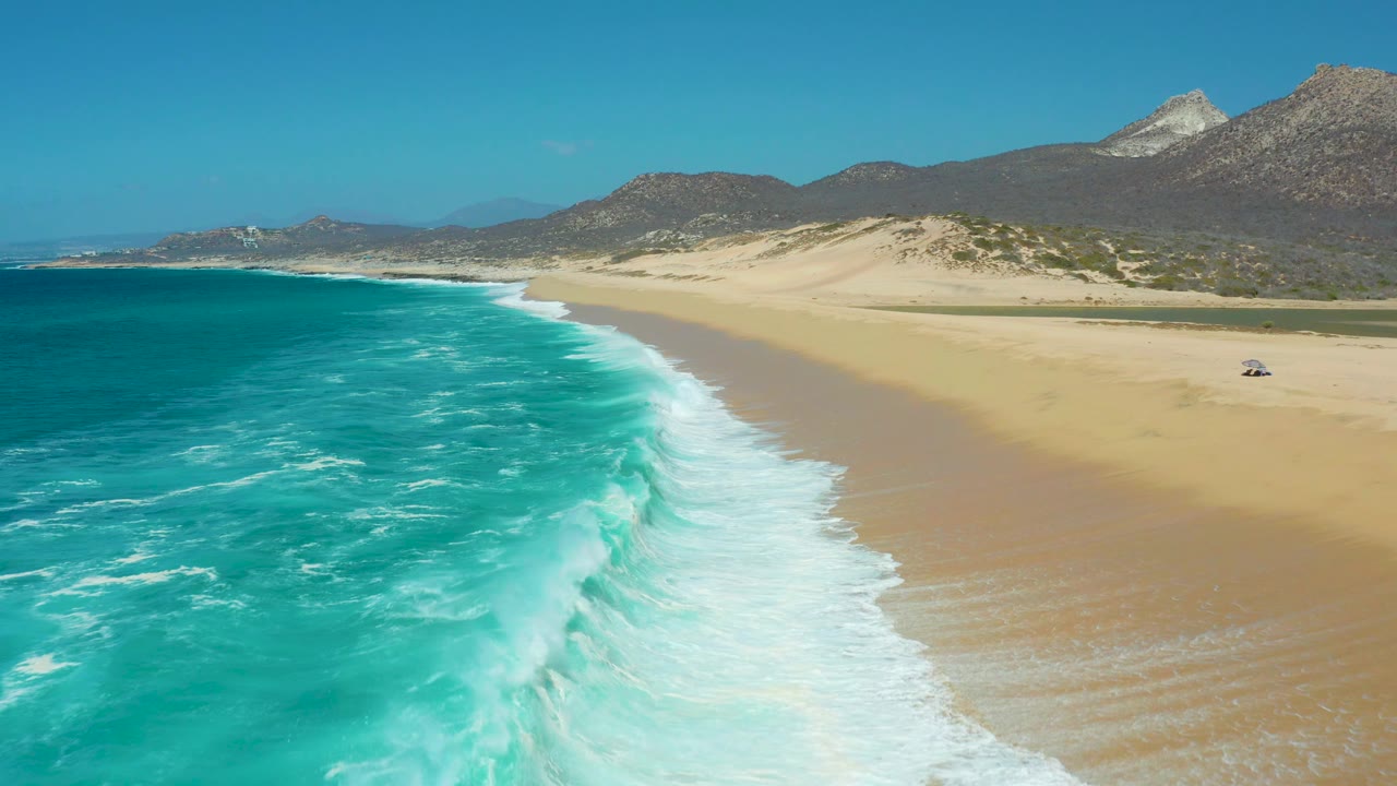 Foamy turquoise waves along a broad beach below dry hills.
