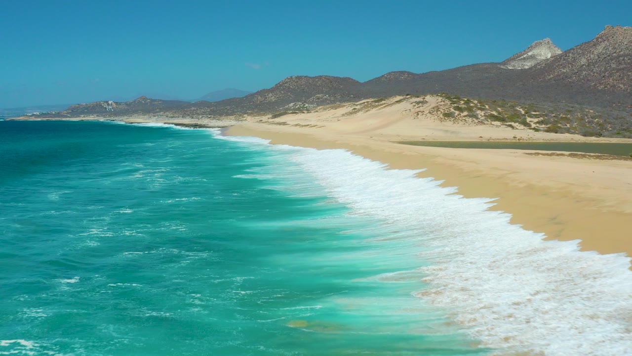 Wide elevated view of La Laguna Beach Los Cabos with turquoise water on the left, white surf reaching pale sand, mountains in the background, and no visible people.