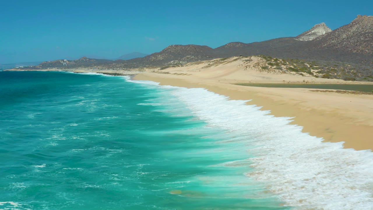 Aerial coastal view at La Laguna Beach Los Cabos with foamy waves spreading farther along the beach, open sand to the right, distant mountains, and no visible people.