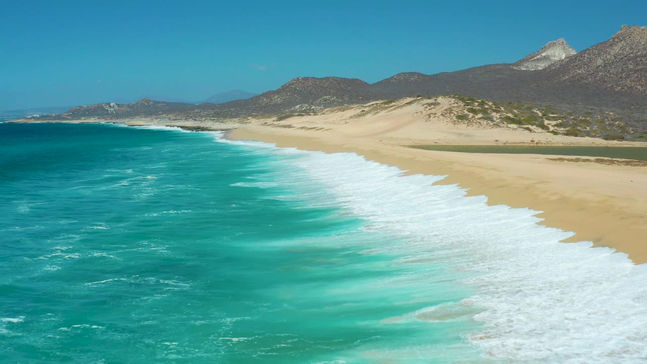 La Laguna Beach Los Cabos shoreline seen from above, with the surf line cutting across the frame, sandy dunes beyond it, dark mountain slopes behind, and no visible people.