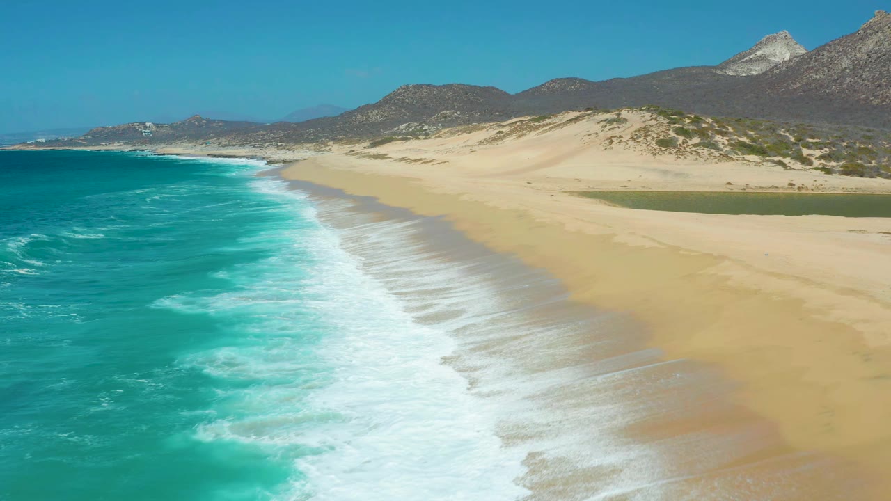 La Laguna Beach aerial view with the surf band slightly farther along the shore, pale sand widening to the right, the lagoon visible near the dunes, and no visible people.