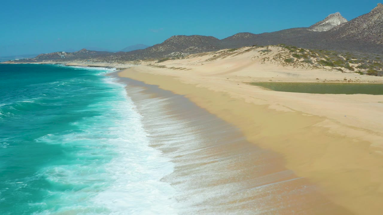 Aerial view at La Laguna Beach with turquoise water on the left, a long diagonal band of foam, smooth sand on the right, dunes and mountains behind, and no visible people.
