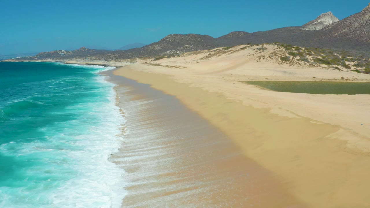 Mid-clip aerial frame of La Laguna Beach showing white water pushing up the left foreground, a broad empty sand flat, a green lagoon near the dunes, mountains, and no visible people.
