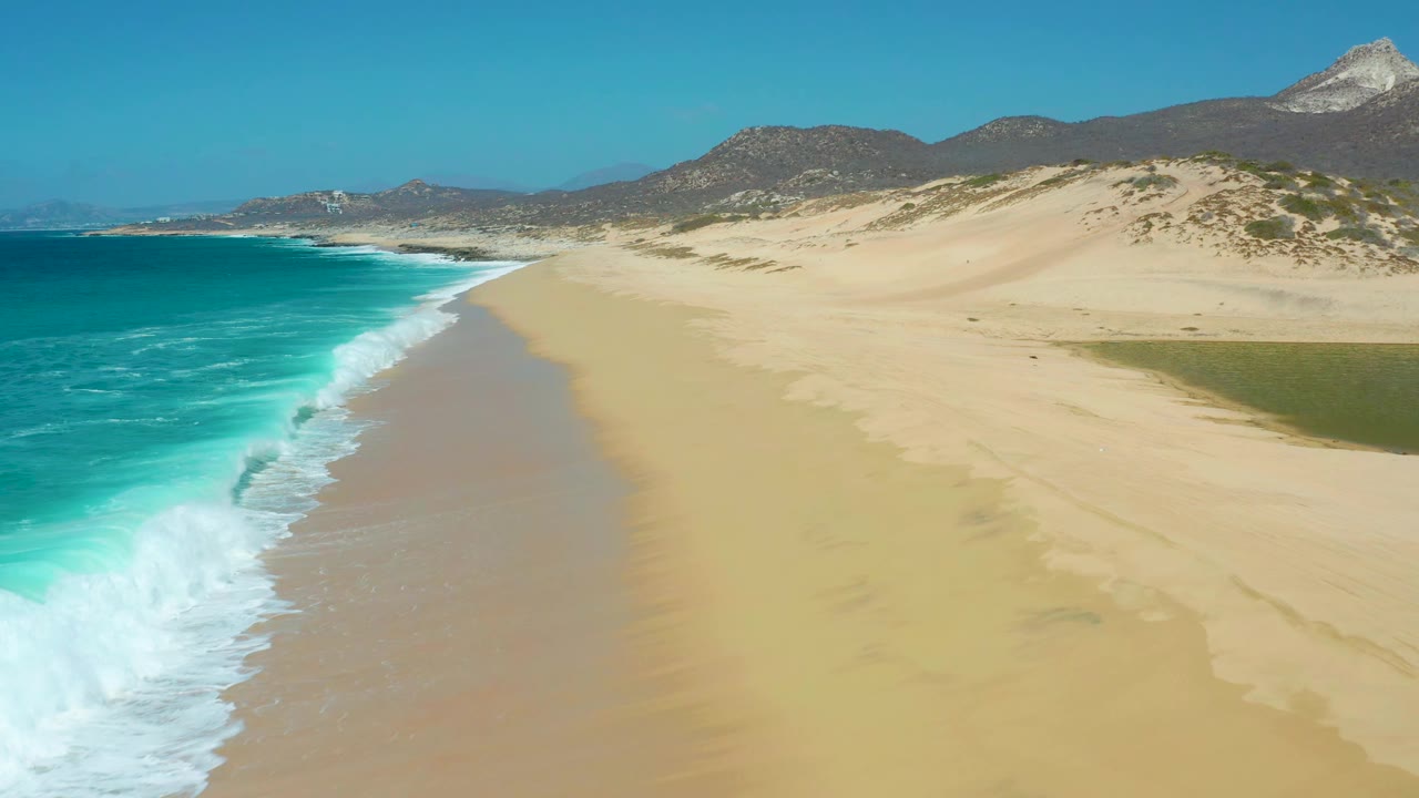 La Laguna Beach Los Cabos shoreline seen from above, with a breaking wave line on the left, open tan sand on the right, distant dune ridges, mountains, and no visible people.