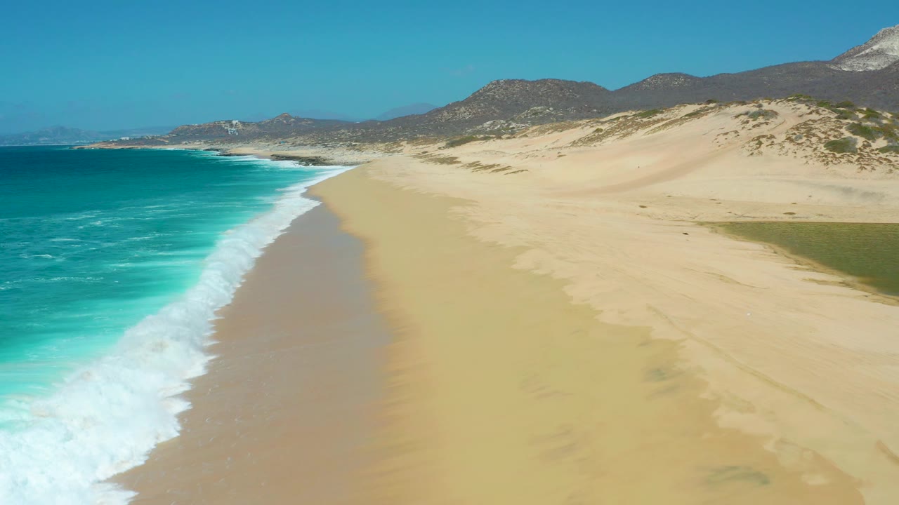 Aerial view at La Laguna Beach Los Cabos with the surf pushed farther into the lower left, a long open beach curving ahead, dune hills to the right, mountains on the horizon, and no visible people.