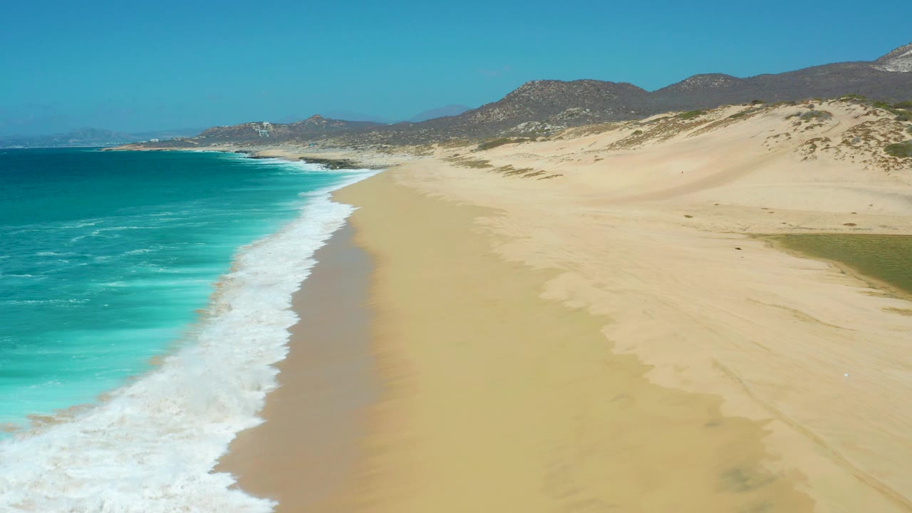 Drone view looking along an empty beach with surf on the left and dunes on the right.