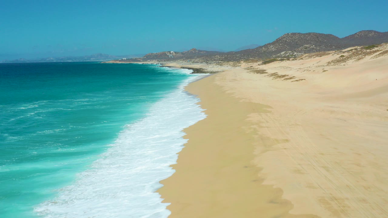 Aerial view of La Laguna Beach Los Cabos, Mexico, with turquoise water on the left, white foam curving onto broad sand, low hills beyond the dunes, and no visible people.