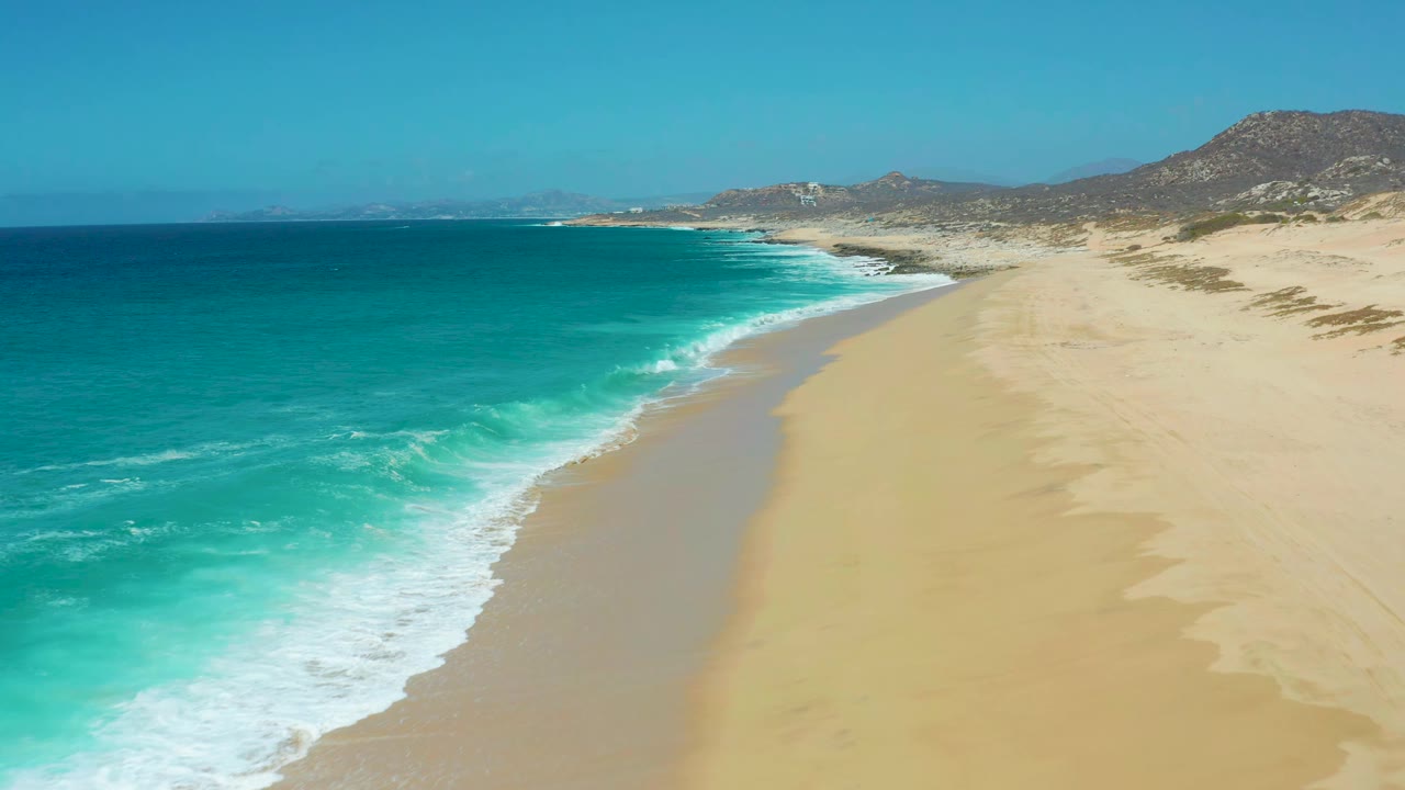 Aerial frame at La Laguna Beach Los Cabos with a stronger band of white foam reaching the wet sand, open turquoise water to the left, dunes to the right, and no visible people.