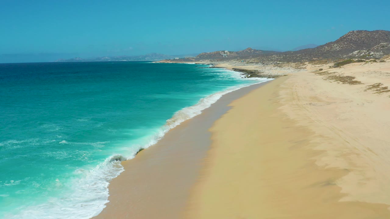 Aerial view along La Laguna Beach Los Cabos where turquoise water breaks into white foam near the lower left, the sandy beach stretches into the distance, and no visible people appear.