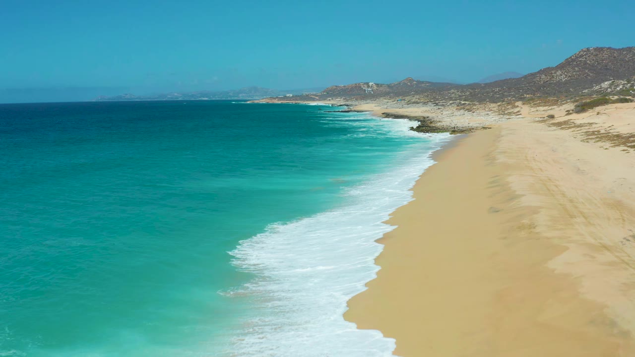 Aerial frame at La Laguna Beach Los Cabos showing a broad band of white surf pushing farther along the sand, with teal water on the left, open beach on the right, hills beyond, and no visible people.