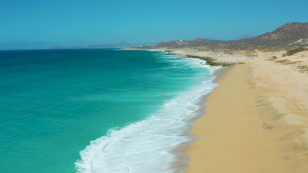 Mid-clip aerial frame over La Laguna Beach Los Cabos, with foamy surf entering from the lower left, a long empty beach on the right, teal water across the left half, distant hills, and no visible people.