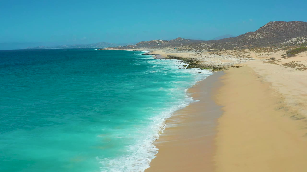 La Laguna Beach coastline seen from above, with the surf line pushed farther down the sand, open turquoise water, rocky coastal ground, and no visible people.