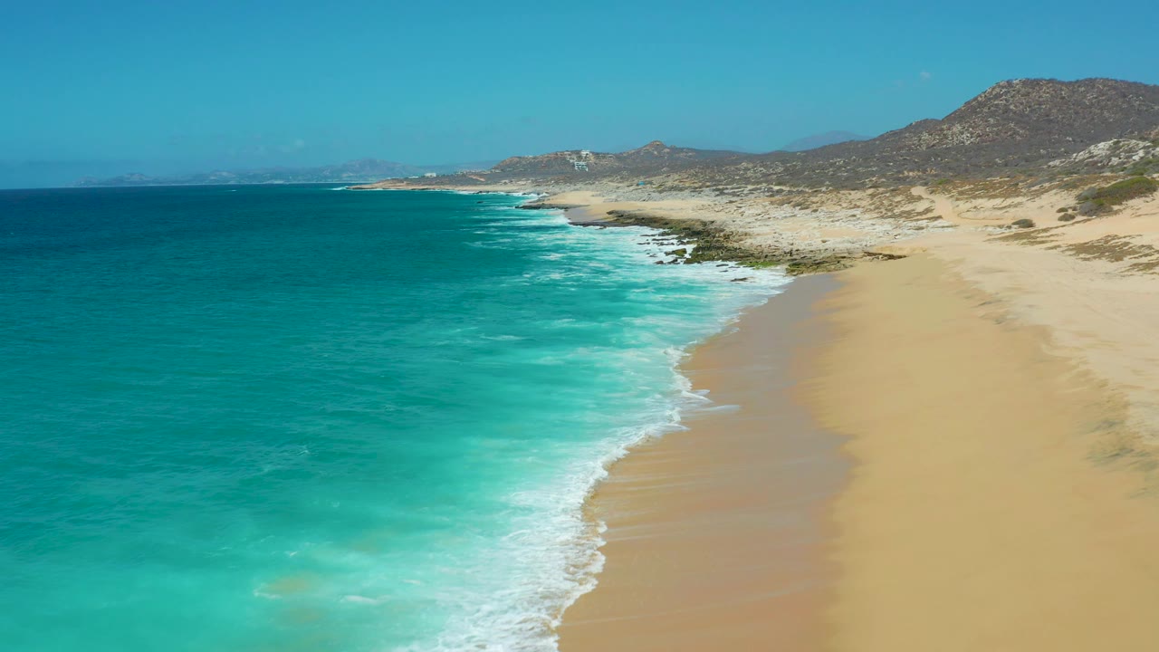 Aerial view over La Laguna Beach with blue-green ocean spreading across the left frame, foam curling onto empty sand, rugged hills behind, and no visible people.