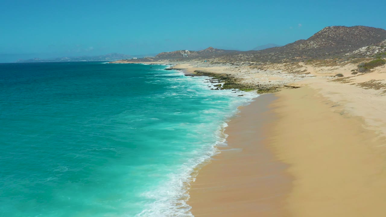 La Laguna Beach from an aerial angle, with turquoise surf advancing across the lower left, a broad empty sand strip on the right, desert-toned hills, and no visible people.