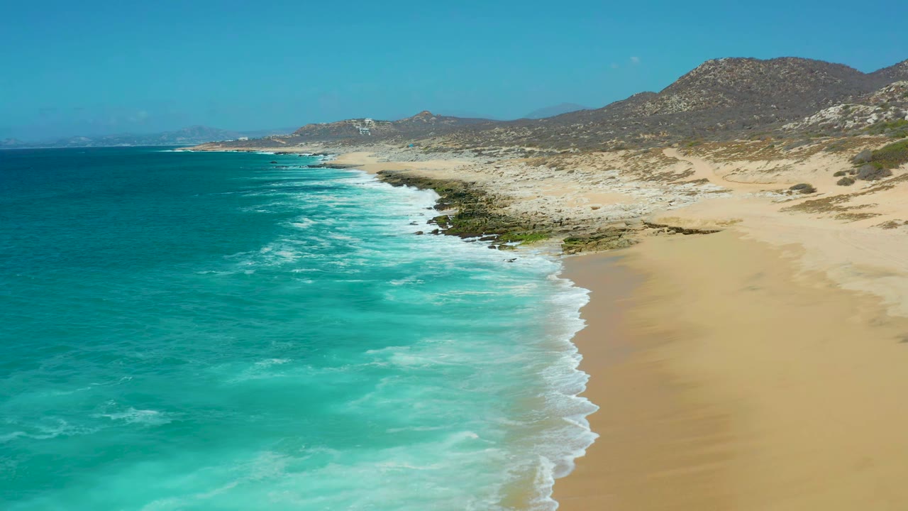 Aerial view at La Laguna Beach in Los Cabos, Mexico, with white foam bands advancing toward a wide tan beach, rocks along the waterline, low hills beyond the sand, and no visible people.