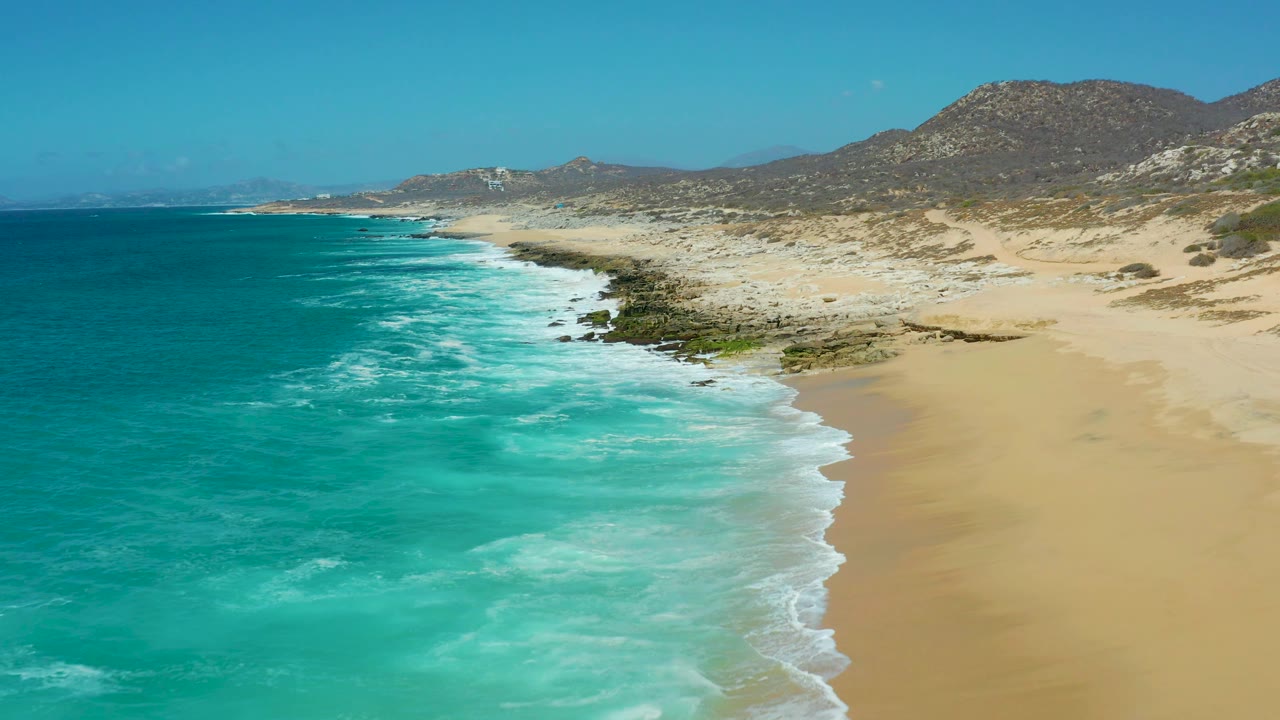 La Laguna Beach aerial frame showing a curved line of white surf, deep turquoise water to the left, open sand to the right, rocky patches near the shore, distant arid hills, and no visible people.