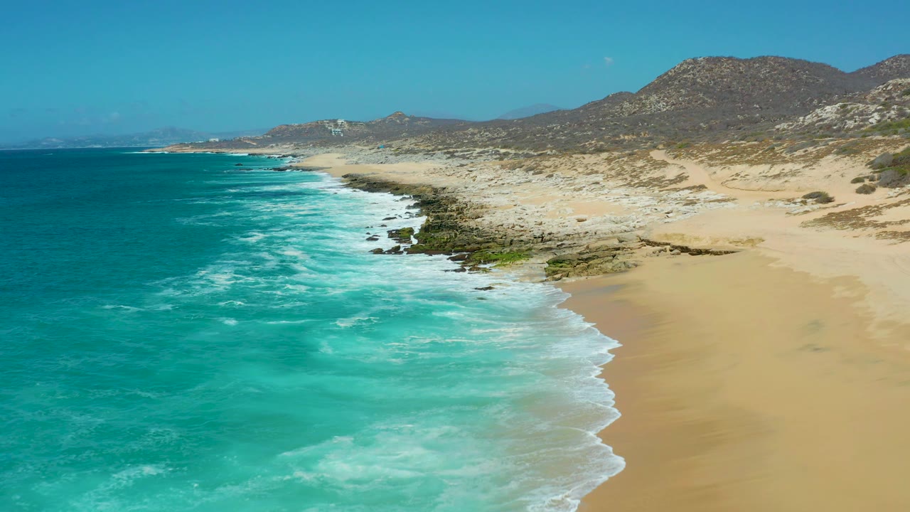 Aerial coastline with surf washing along sand and rocks.