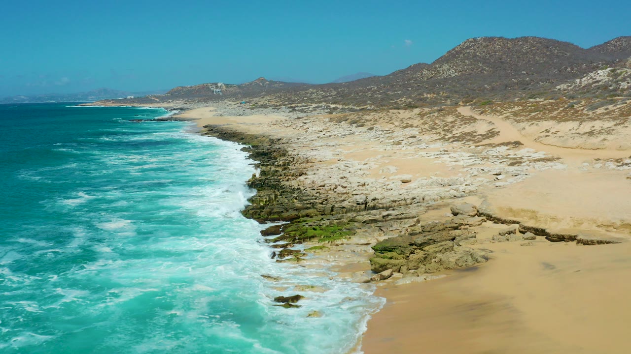 Aerial view over La Laguna Beach Los Cabos with turquoise ocean on the left, foam hitting rocks and sand on the right, dry hills behind, and no visible people.