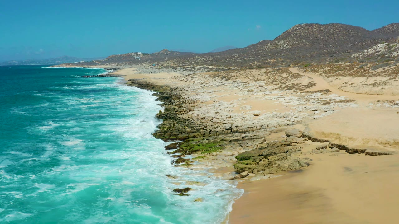 Aerial frame at La Laguna Beach Los Cabos showing a wider run of turquoise water, white foam bands, rocky outcrops, pale sand, arid hills, and no visible people.