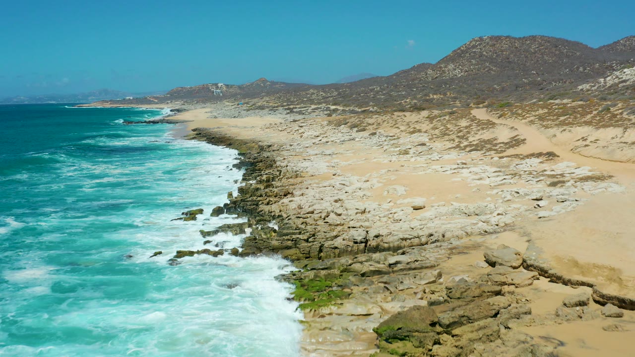 Surf line beside a rocky beach with sand dunes and low mountains.