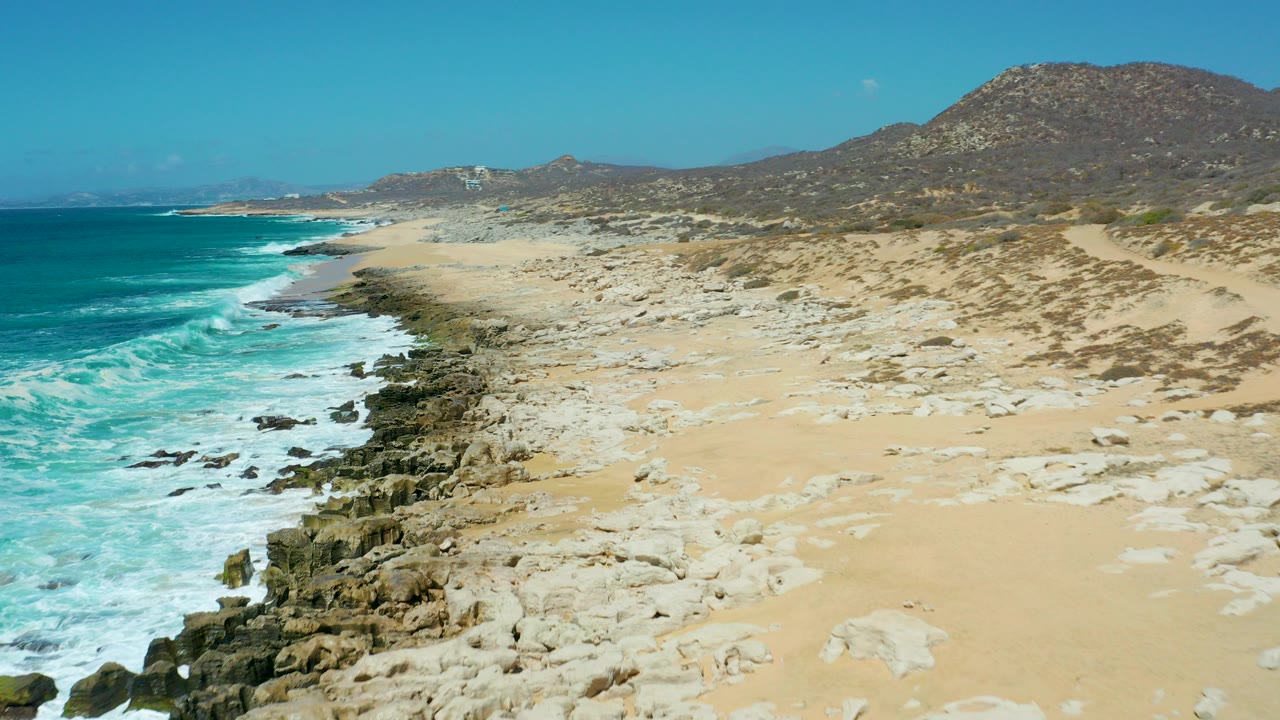 La Laguna Beach in Los Cabos with turquoise water on the left, pale sand and rock shelves across the shore, dry hills in the distance, and no visible people.