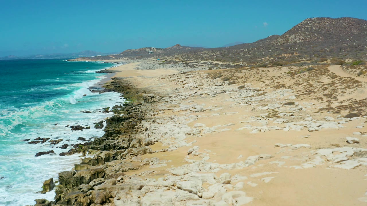 A wide La Laguna Beach view shows the rocky shoreline curving through the frame, turquoise water to the left, open sand to the right, hills beyond, and no visible people.