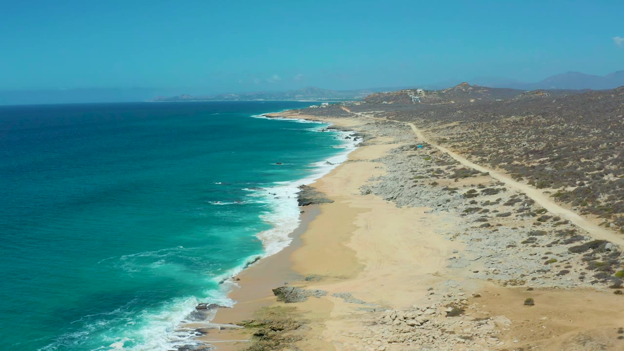 Aerial view at La Laguna Beach Los Cabos with white foam tracing the shoreline, pale sand below, dry brushland beside the dirt road, distant hills, and no visible people.