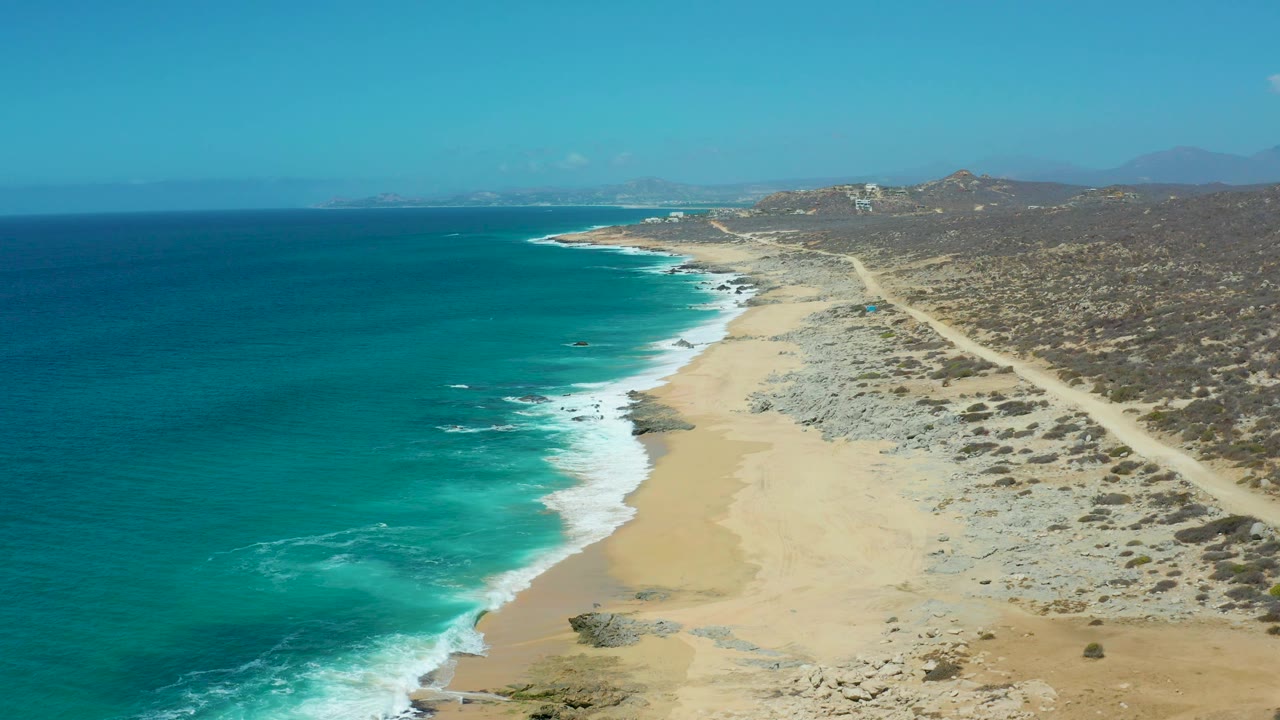 Wide aerial frame of La Laguna Beach Los Cabos showing a broader stretch of turquoise water, breaking waves, a sandy shoreline, a pale road inland, scrub-covered terrain, and no visible people.