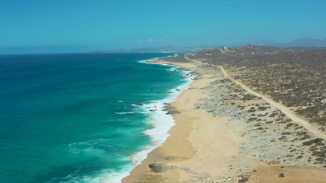 La Laguna Beach Los Cabos aerial frame with deep blue water at far left, bright turquoise shallows near shore, white surf against sand and rocks, road and scrubland on the right, and no visible people.