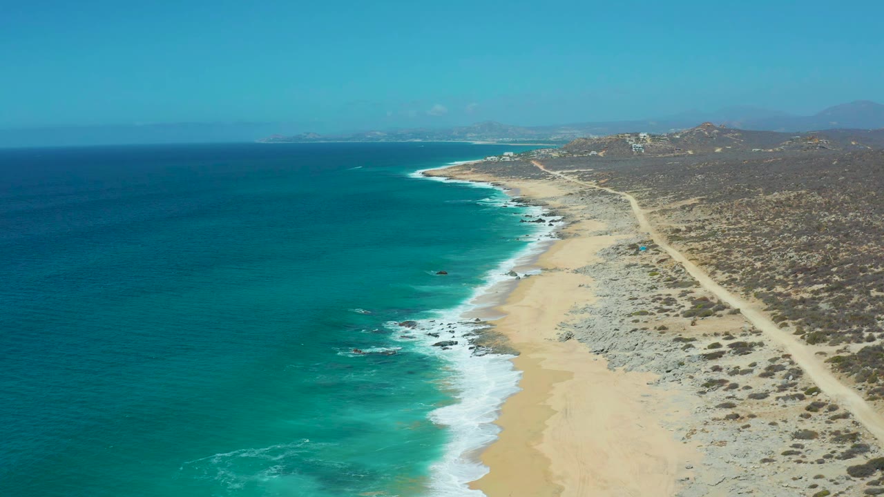 Aerial coastline with blue-green water, surf, sand, rocks, and arid land.