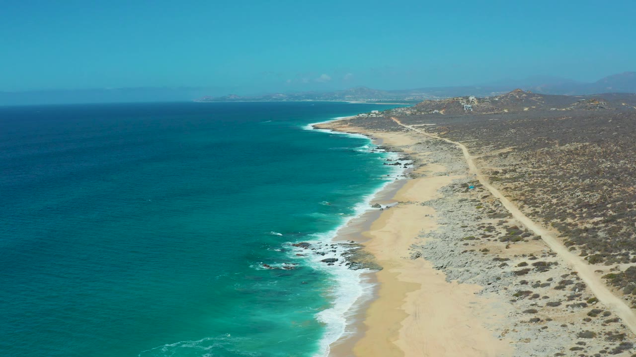 Drone view over ocean and sandy beach with rocky patches and dry roadside terrain.