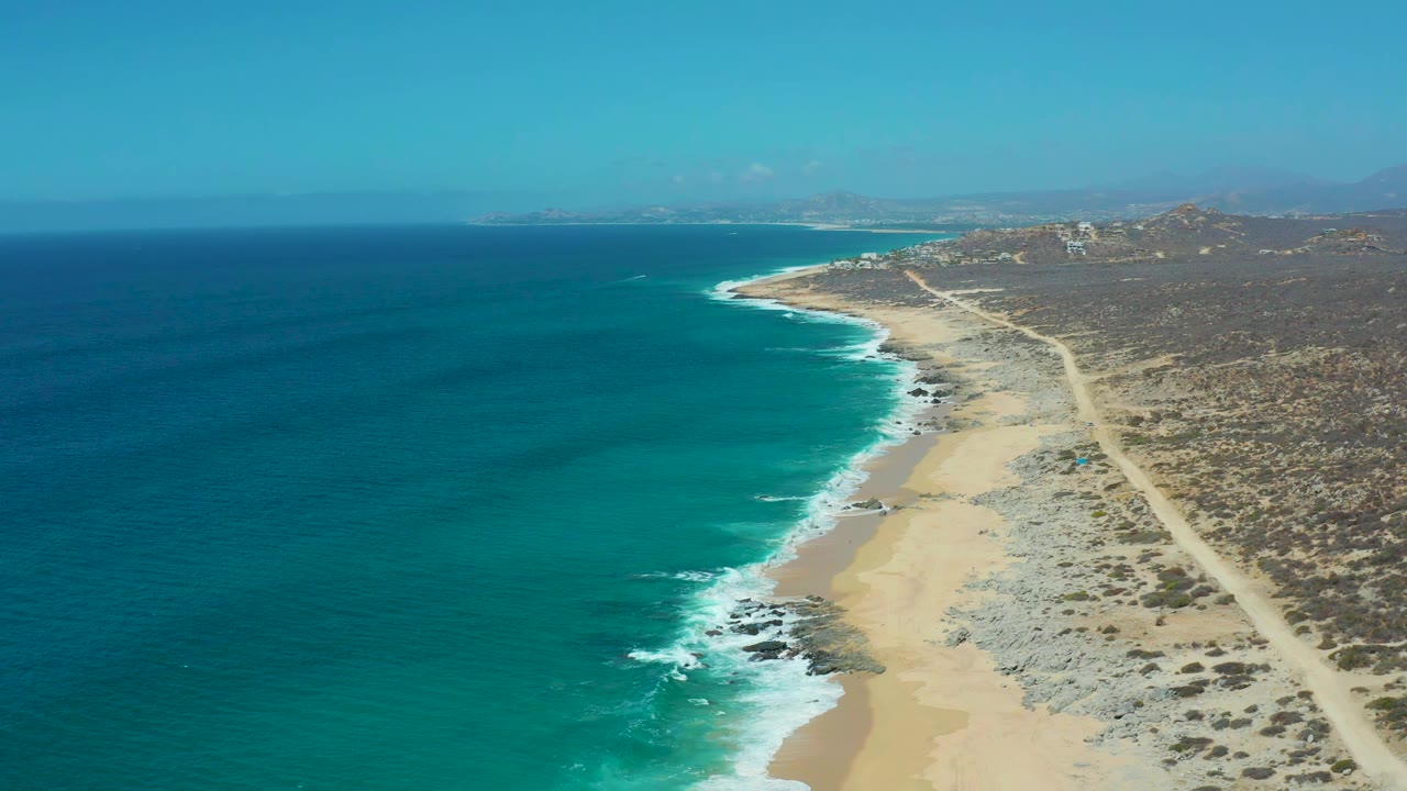 Aerial frame of La Laguna Beach in Los Cabos, Mexico showing deep blue water grading to turquoise near shore, white wave lines, pale sand, a dirt road through arid terrain, and no visible people.