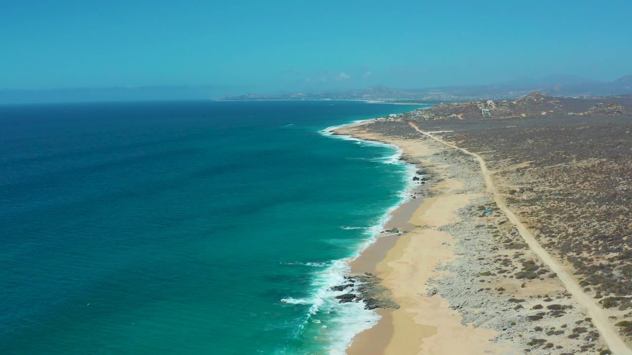 A high aerial view at La Laguna Beach in Los Cabos, Mexico shows the ocean occupying most of the left frame, white surf reaching rocky sand, a straight dirt road inland, and no visible people.