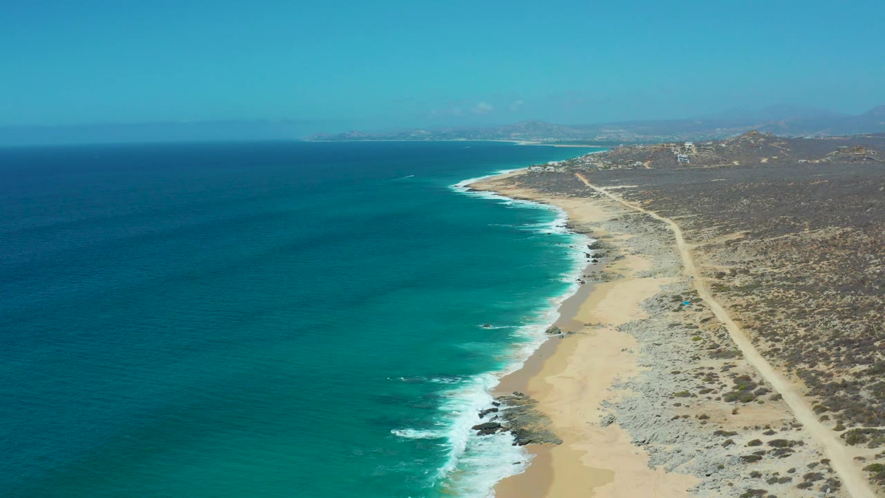 Mid-clip aerial over La Laguna Beach in Los Cabos, Mexico with blue water, breaking surf, sandy and rocky shoreline, a dirt road beyond the beach, hazy distant hills, and no visible people.
