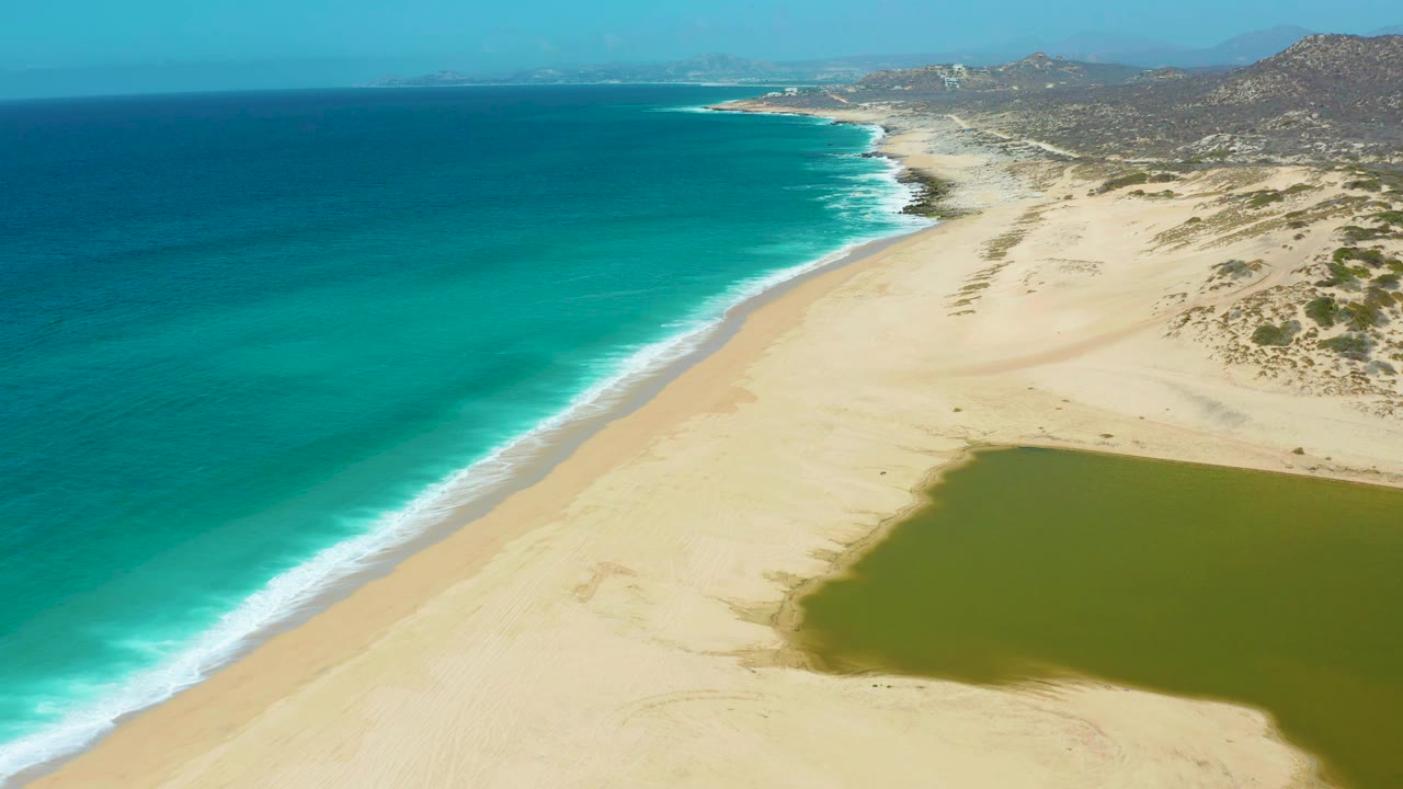 Aerial frame at La Laguna Beach Los Cabos, Mexico, with the surf line running diagonally beside wide sand, the green lagoon occupying the lower right, and no visible people.