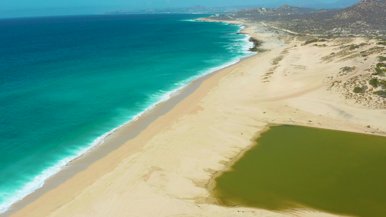 Aerial frame over La Laguna Beach Los Cabos, Mexico, with turquoise ocean left, a narrow white wave edge, pale beach center, lagoon corner at lower right, distant rocky hills, and no visible people.