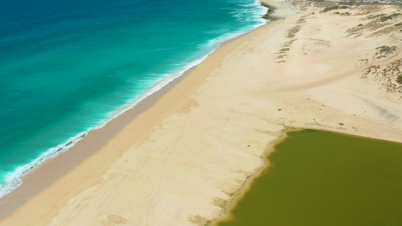 Aerial view of turquoise ocean, sandy beach, dunes, and green lagoon.