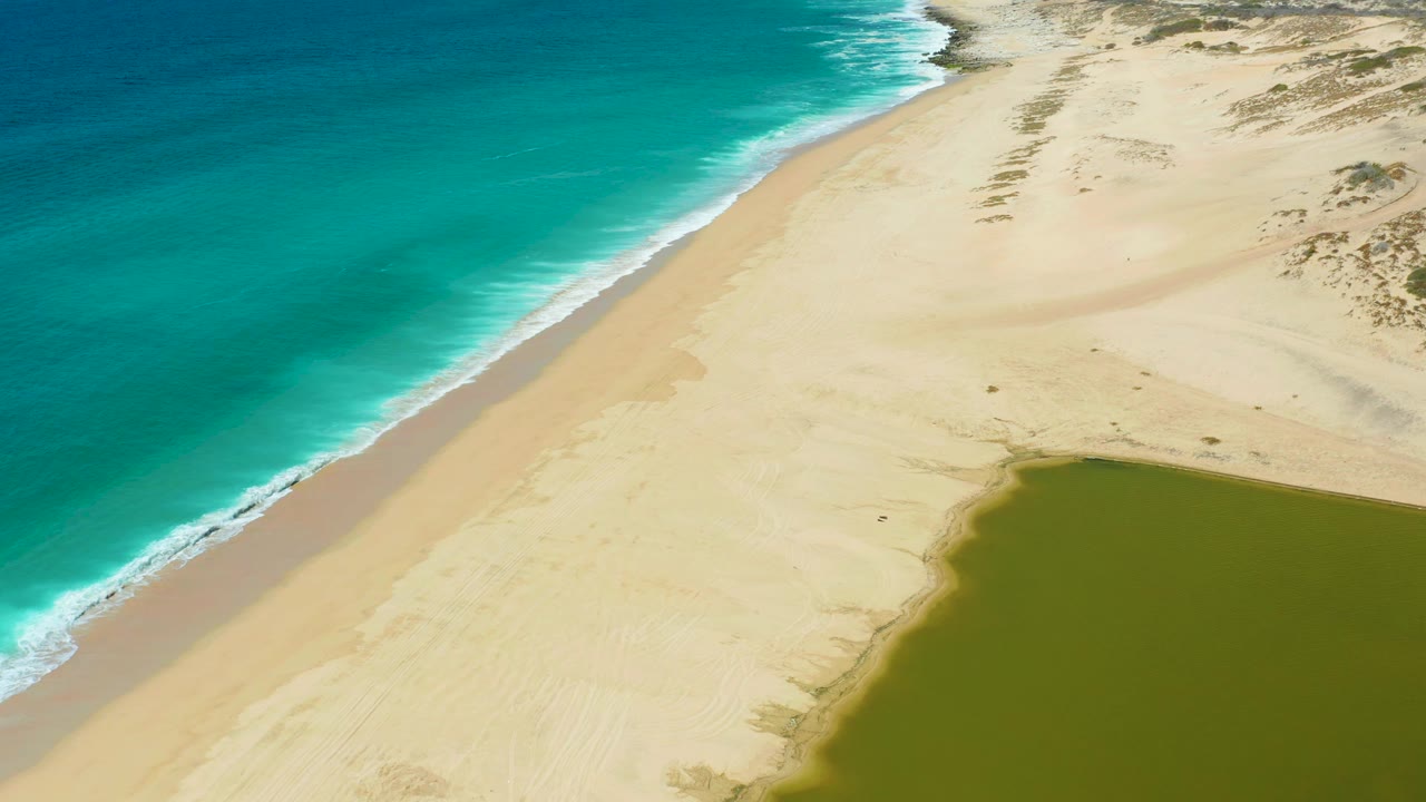 Aerial view over La Laguna Beach with the surf line running diagonally, pale sand widening inland, the lagoon edge entering the lower right, and no visible people.