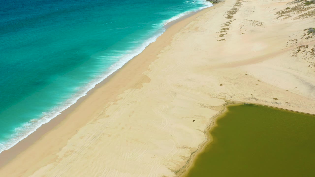 Aerial view of La Laguna Beach with more surf visible along the left edge, open sand across the middle, sparse dune vegetation above, green lagoon at lower right, and no visible people.